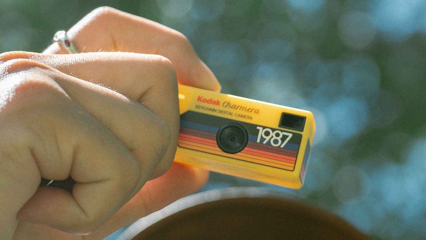 A hand holding up a yellow coloured Kodak Charmera with the sky and a tree behind him