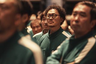 A man in glasses (Yang Dong-geun) stands amid a crowd of people, blurred around him, all wearing matching green tracksuits, in 'Squid Game' season 2.