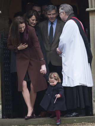 Kate Middleton wearing a burgundy coat and heels holding hands with a young Princess Charlotte who is holding a candy cane and walking out of church with Carole and Michael Middleton and a minister behind them