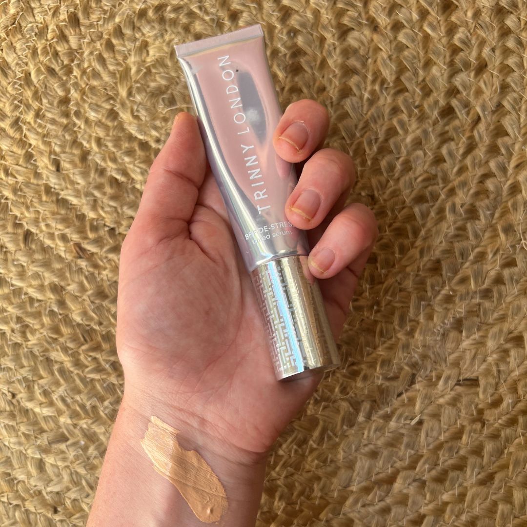 Beauty Editor Fiona McKim's hand holding a tube of Trinny London BFF foundation, with a swatch of the foundation on her wrist, on a woven jute rug backdrop