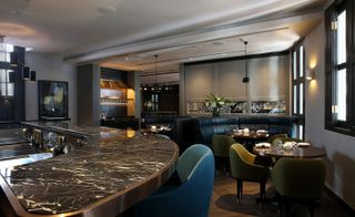Interior view of the bar and a dining area at Disgruntled Chef featuring grey walls, wooden flooring, pendant lights, a curved marble effect bar counter, blue and muted green chairs, wooden tables with tableware and dark coloured curved seating