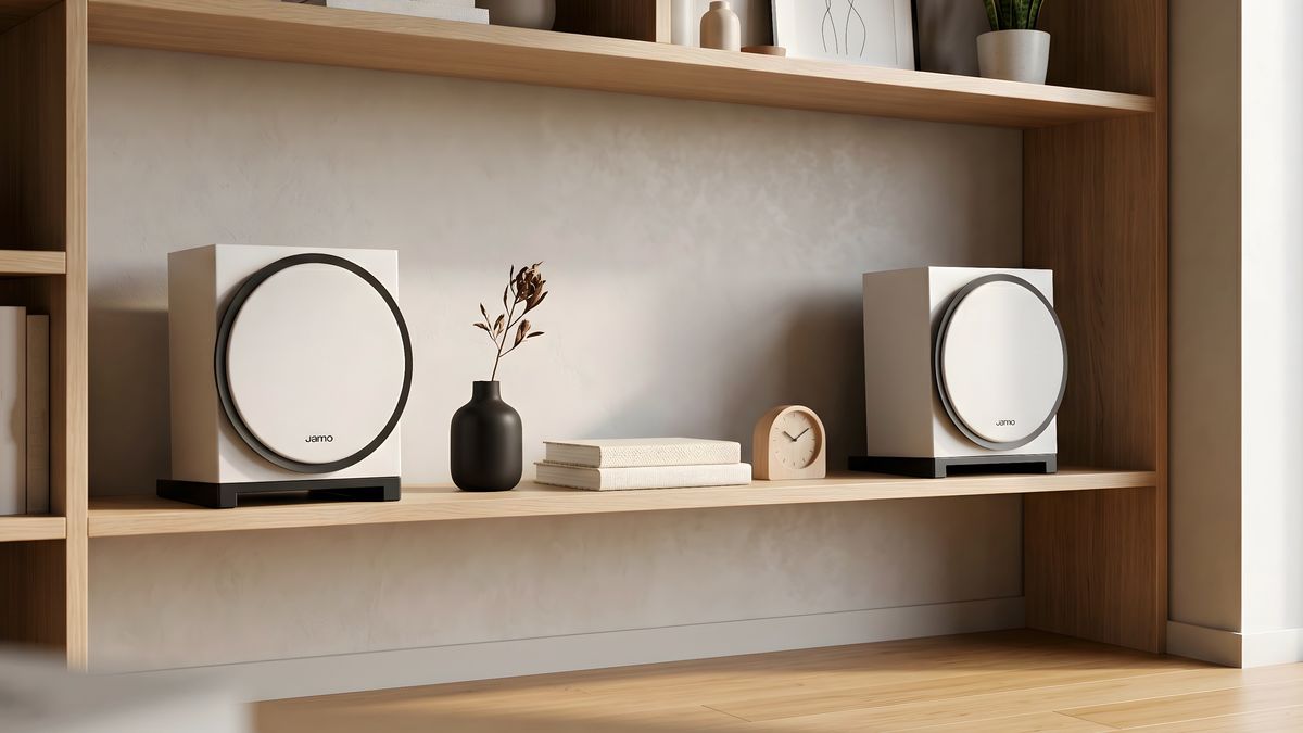 A white pair of Jamo Concert Element 50 speakers on a bookshelf with books, a clock and a diffuser between them.
