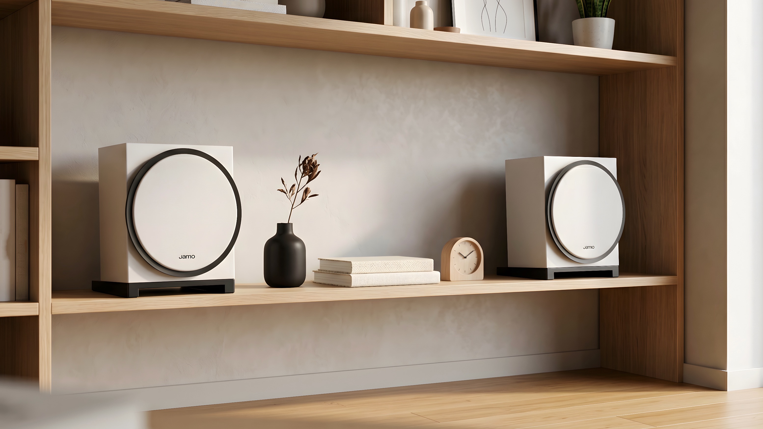 A white pair of Jamo Concert Element 50 speakers on a bookshelf with books, a clock and a diffuser between them.