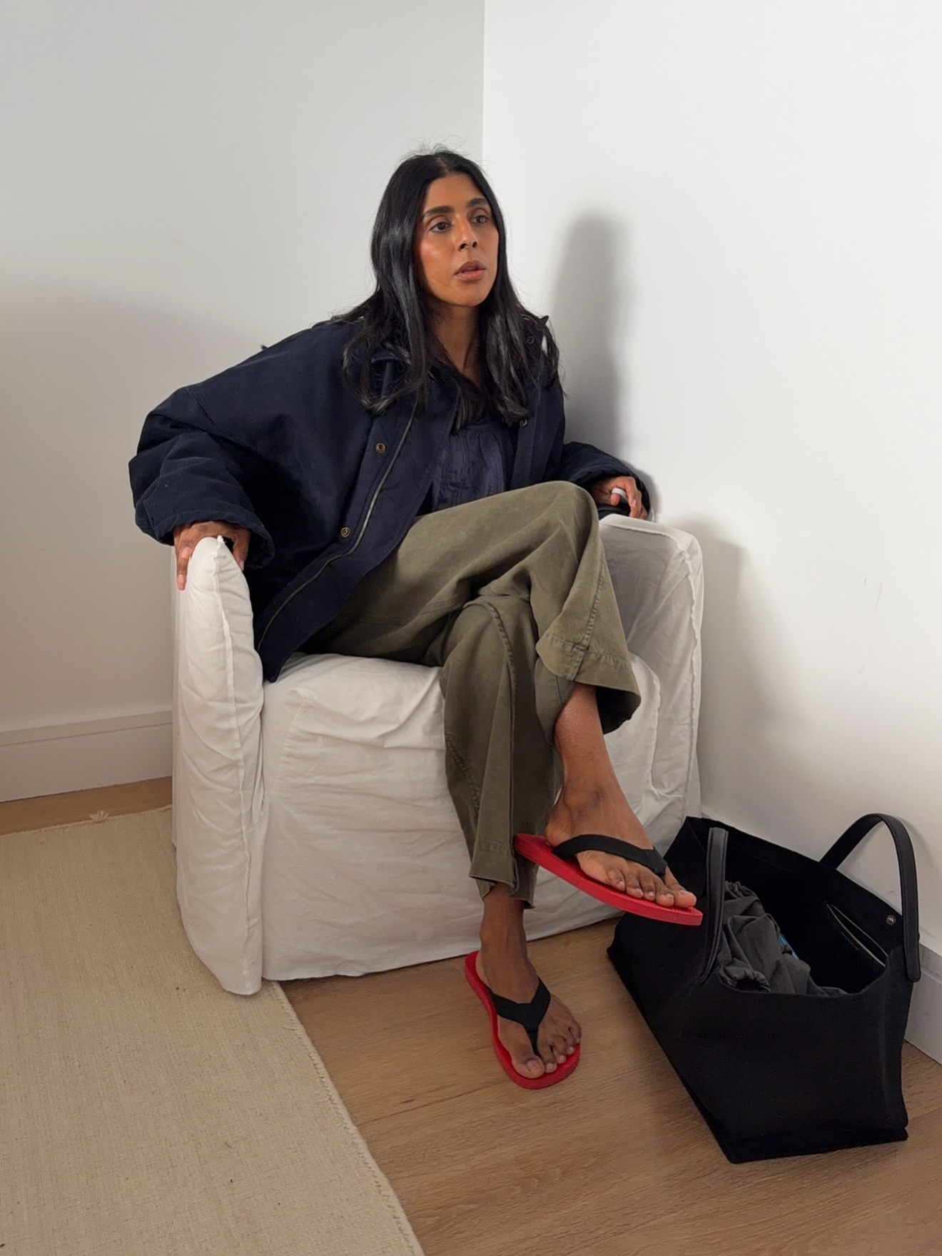 British style influencer Monikh Dale poses in a white slipcover chair wearing an oversize navy blue jacket baggy green pants, black suede tote bag, and black and red flip-flop sandals