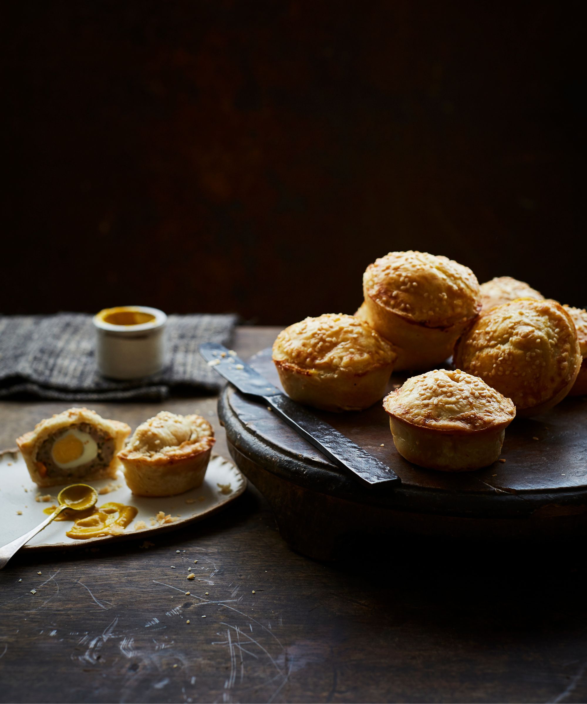 Lamb and egg pies on a round serving board with one pie cut open on a small plate with a spoonful of mustard next to it