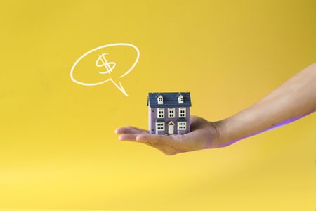 Hand holding a miniature house with a dollar sign speech bubble on yellow background.