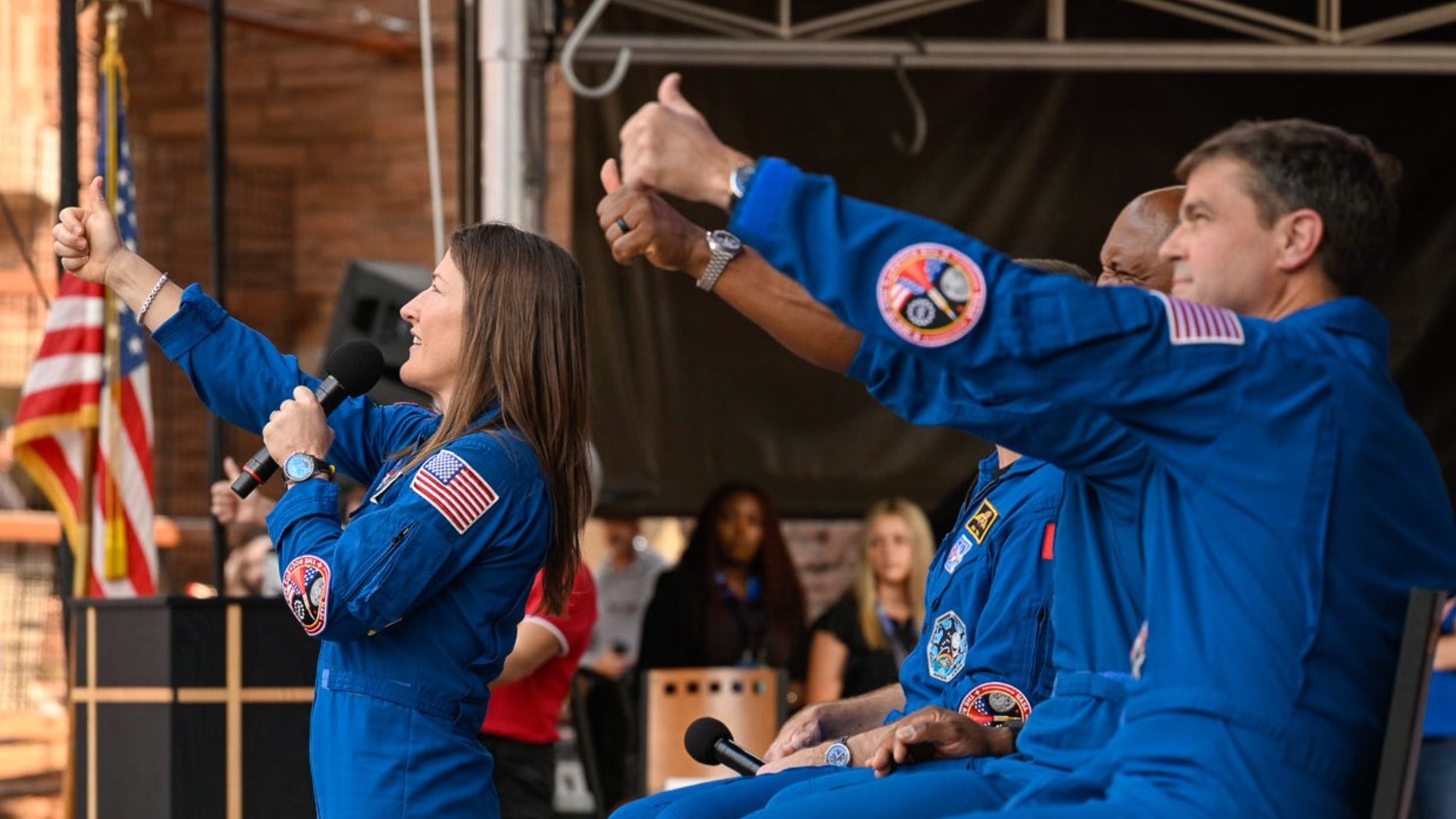 four people in blue flight suits stand on a stage in an amphitheater surrounded by red stone cliffs