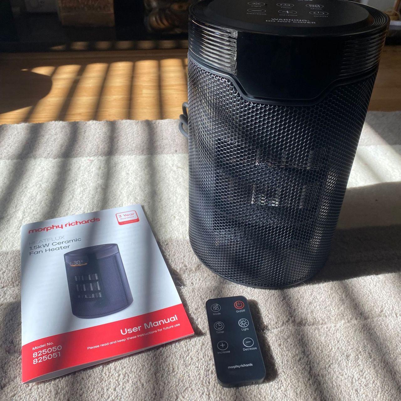 Morphy Richards HeatFlux 1.5kW Ceramic Fan Heater, instruction manual and remote control on neitral rug, with sunlight shining on them