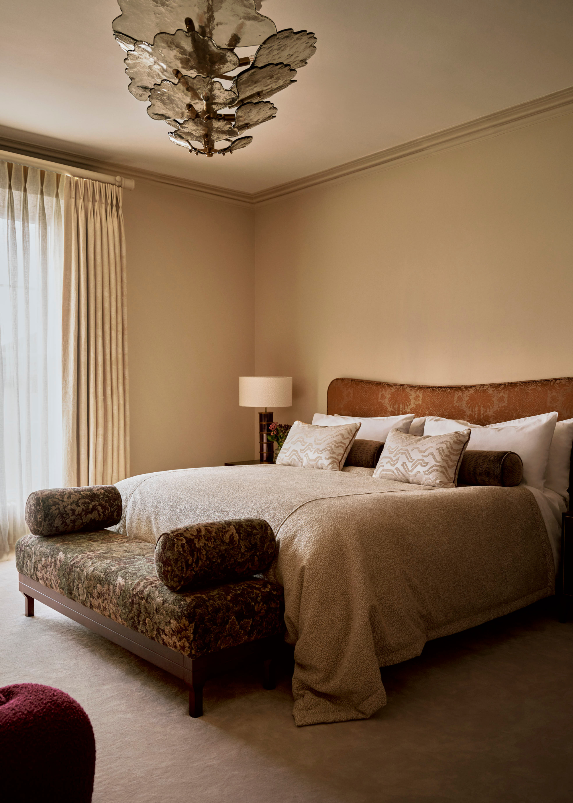 A cream bedroom with a petalled glass ceiling light, a bed with a patterned headboard, fresh pillows, and an animal print bench