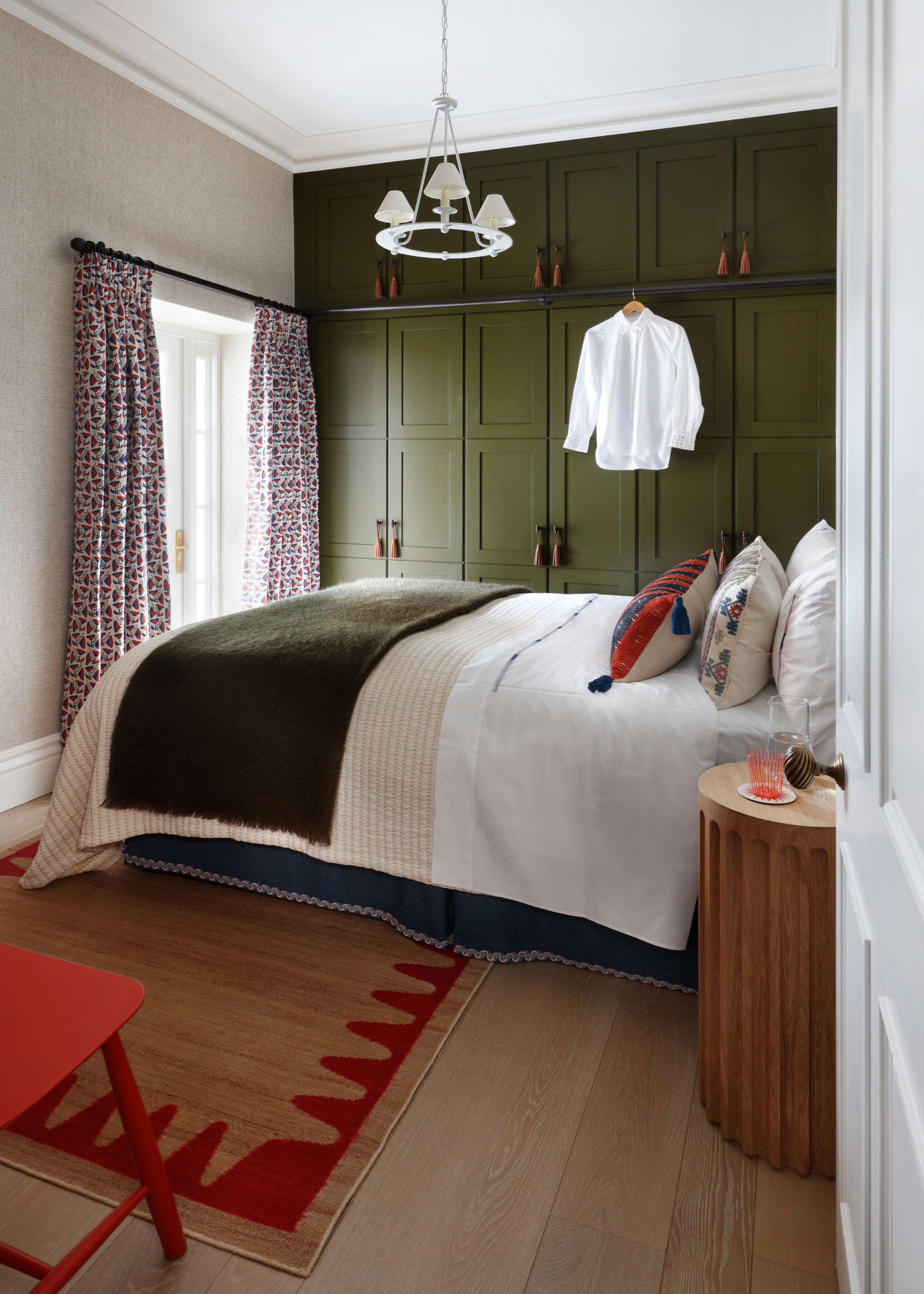 A modern bedroom with white walls and olive green full-height wardrobes. Wood floors with a jute and red rug, neutral bedding with a dark green throw, and patterned curtains.