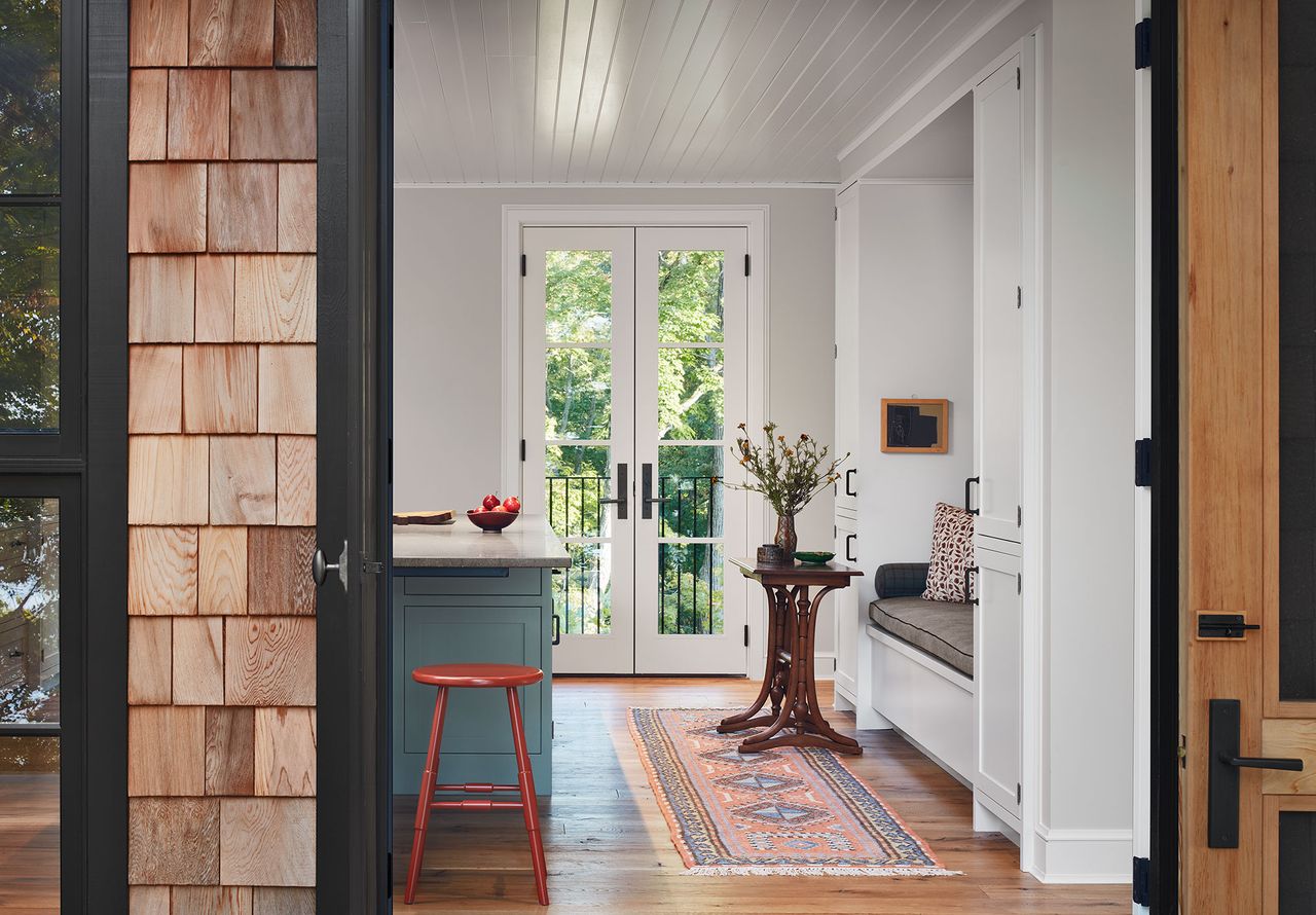 kitchen door with view of blue island white walls and wood paneled ceiling orange stool and banquette with kilim rug and French windows vintage side table with fresh flowers