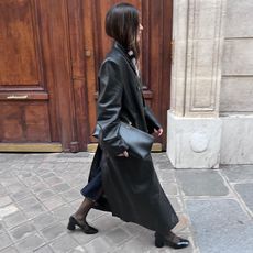 woman wears black trench coat while holding a giant large black clutch and black pumps with tights