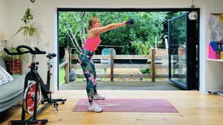 Personal trainer Maddy Biddulph performs a kettlebell swing. She wears a pink vest, tropical-print leggings and white sneakers. She is standing, with her her torso leaning forward at a slight angle, as she swings a kettlebell up in front of her face with her arms straight. Behind her is a leafy garden and nearby is an exercise bike.