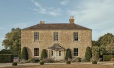 Exterior view of a classic two-story stone country house with symmetrical windows, a central portico, and sculpted garden hedges