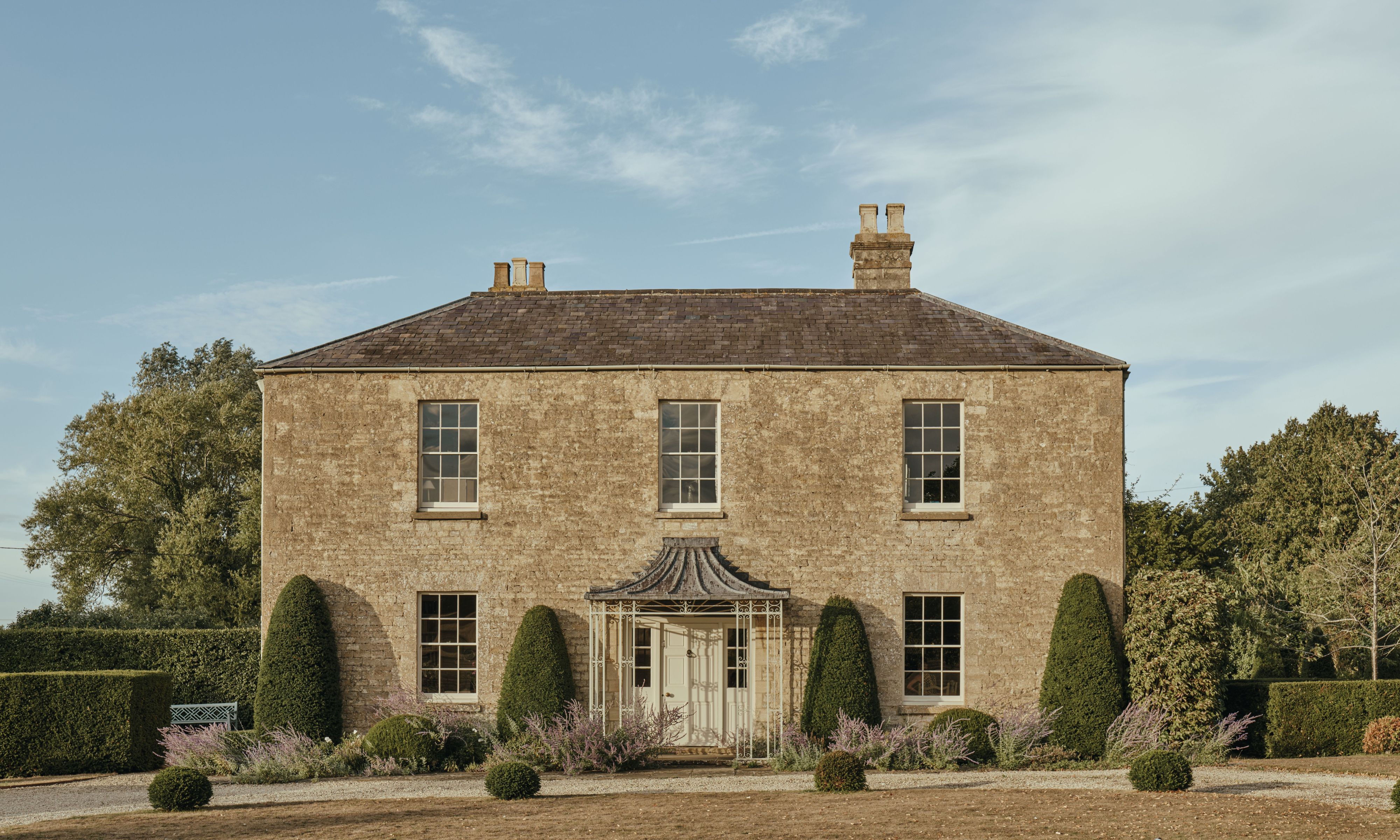 Exterior view of a classic two-story stone country house with symmetrical windows, a central portico, and sculpted garden hedges