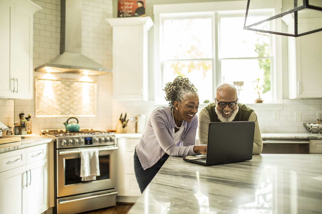 An older couple smiles as they look at their laptop on the kitchen island.