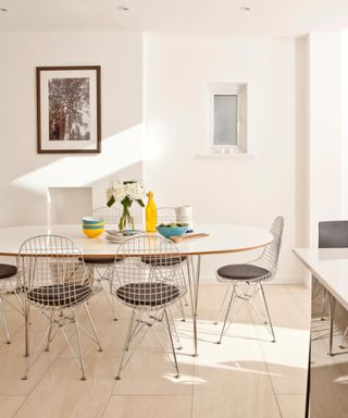 A cream colored dining room with a white wall with two framed wall art prints, a circular white table with yellow and blue bottles and dinnerware, and six metal chairs with black seat covers