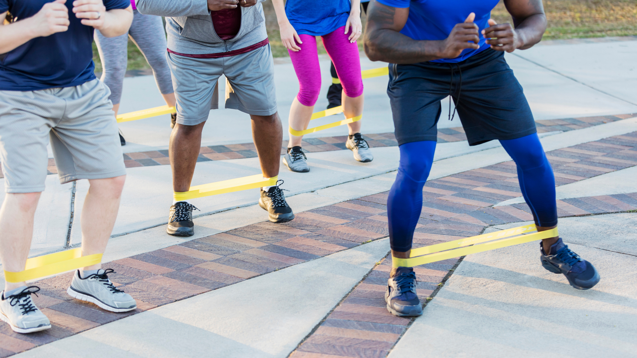 A group of people seen from the waist down, exercising with a small loop resistance band wrapped around their ankles