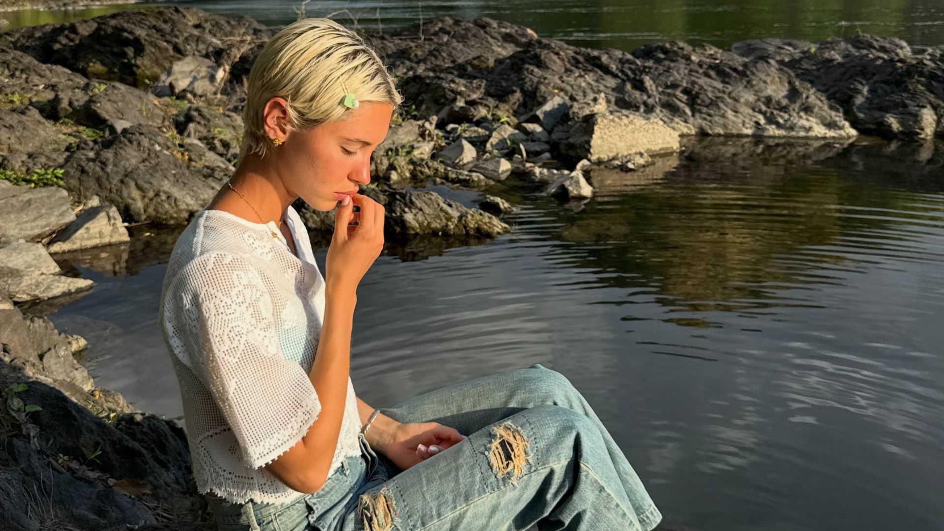 Iris Law sits by a lake wearing ripped jeans with a white boho blouse.