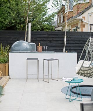 A white built in outdoor bar with stools and a pizza oven in front of a dark fence
