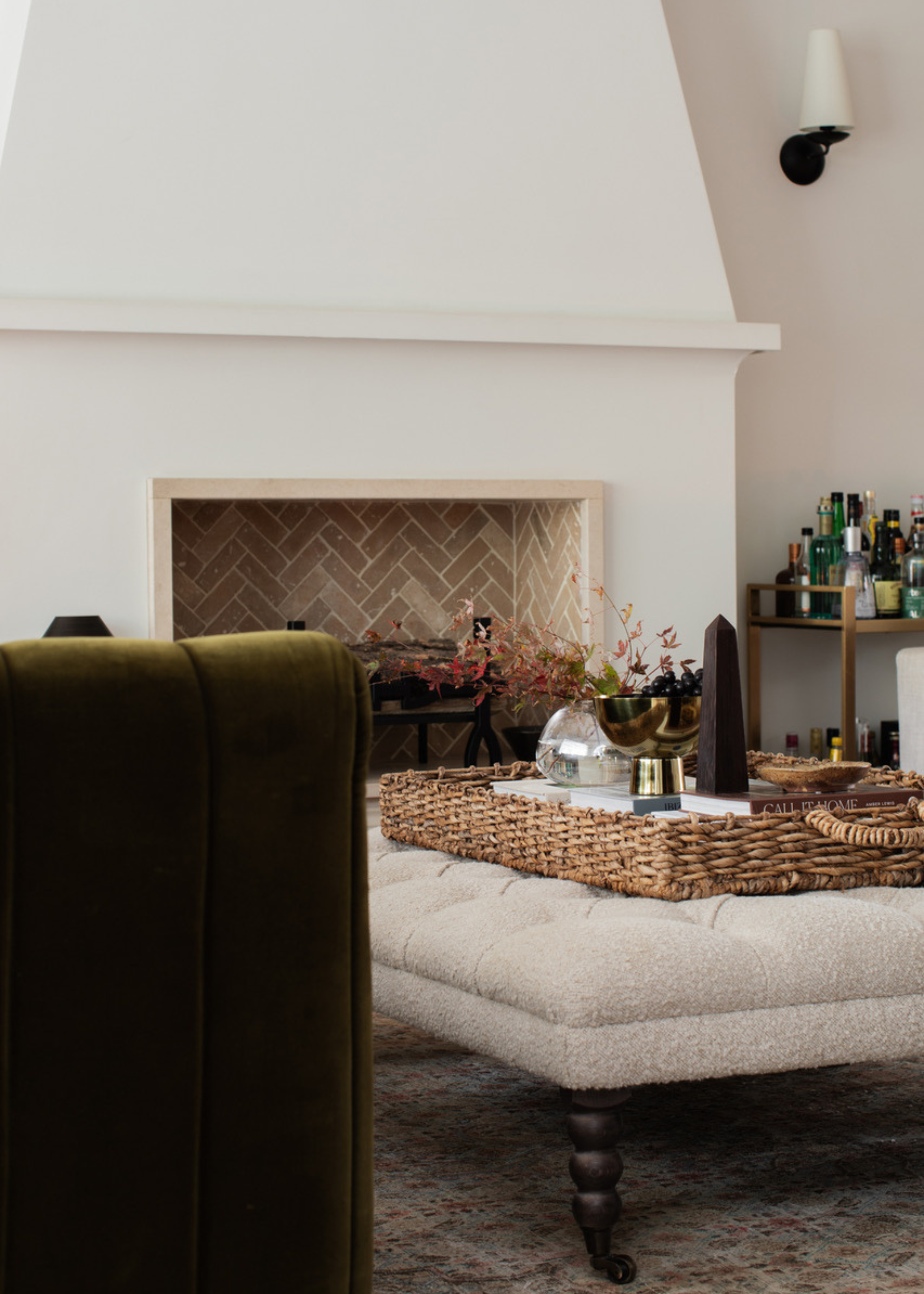 A white living room with a tiled fireplace, a patterned rug, an upholstered coffee table with a woven tray and a bar cart