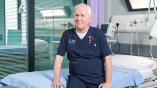 Derek Thompson as Charlie Fairhead in Casualty sitting on a hospital bed and smiling 