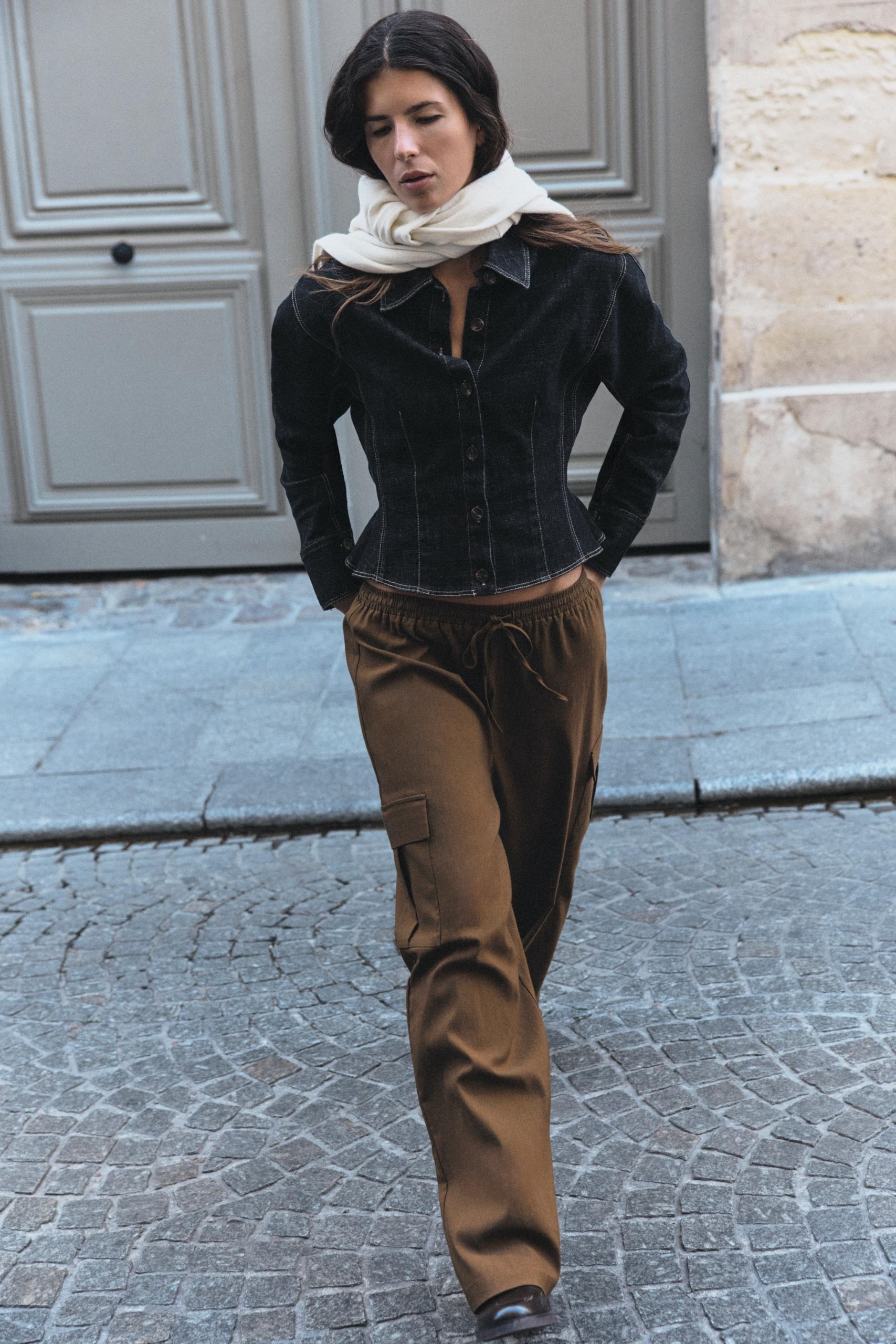 Woman wears peplum denim jacket, cargo pants, white sweater as a scarf, and tall boots.