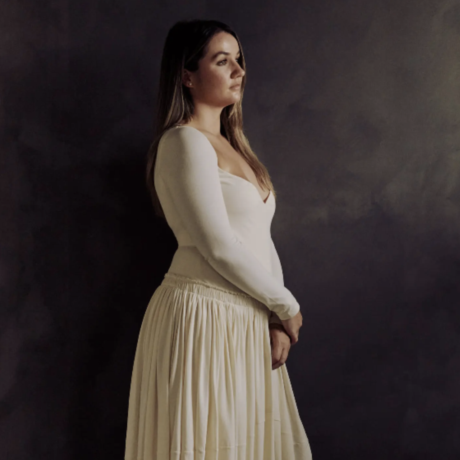A woman in a white flowy dress, leaning against a black wall