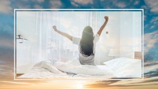 A woman sat on edge of white bed stretching her arms overhead looking out to morning sun