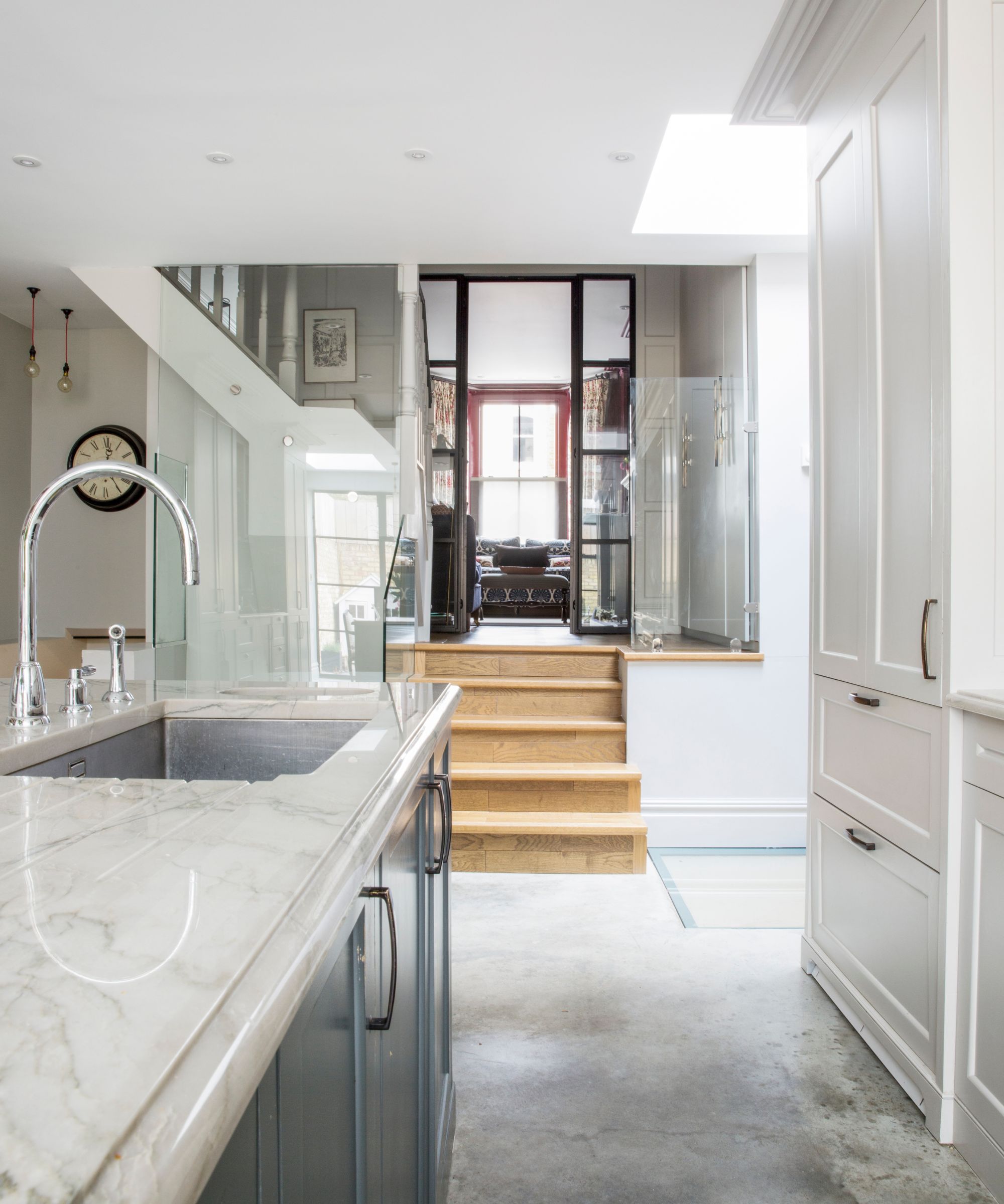 A kitchen sink with marble countertops, blue cabinet, white cabinets, and a wooden staircase down into the kitchen.