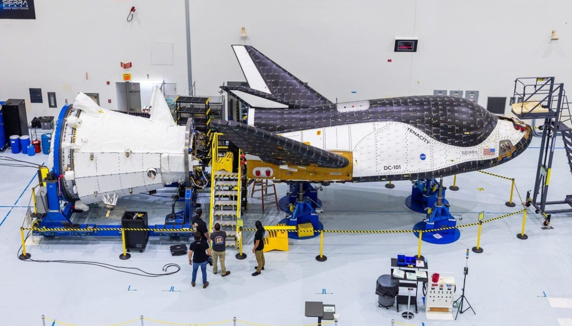 A large black and white space shuttle gets worked on by crews inside a clean warehouse