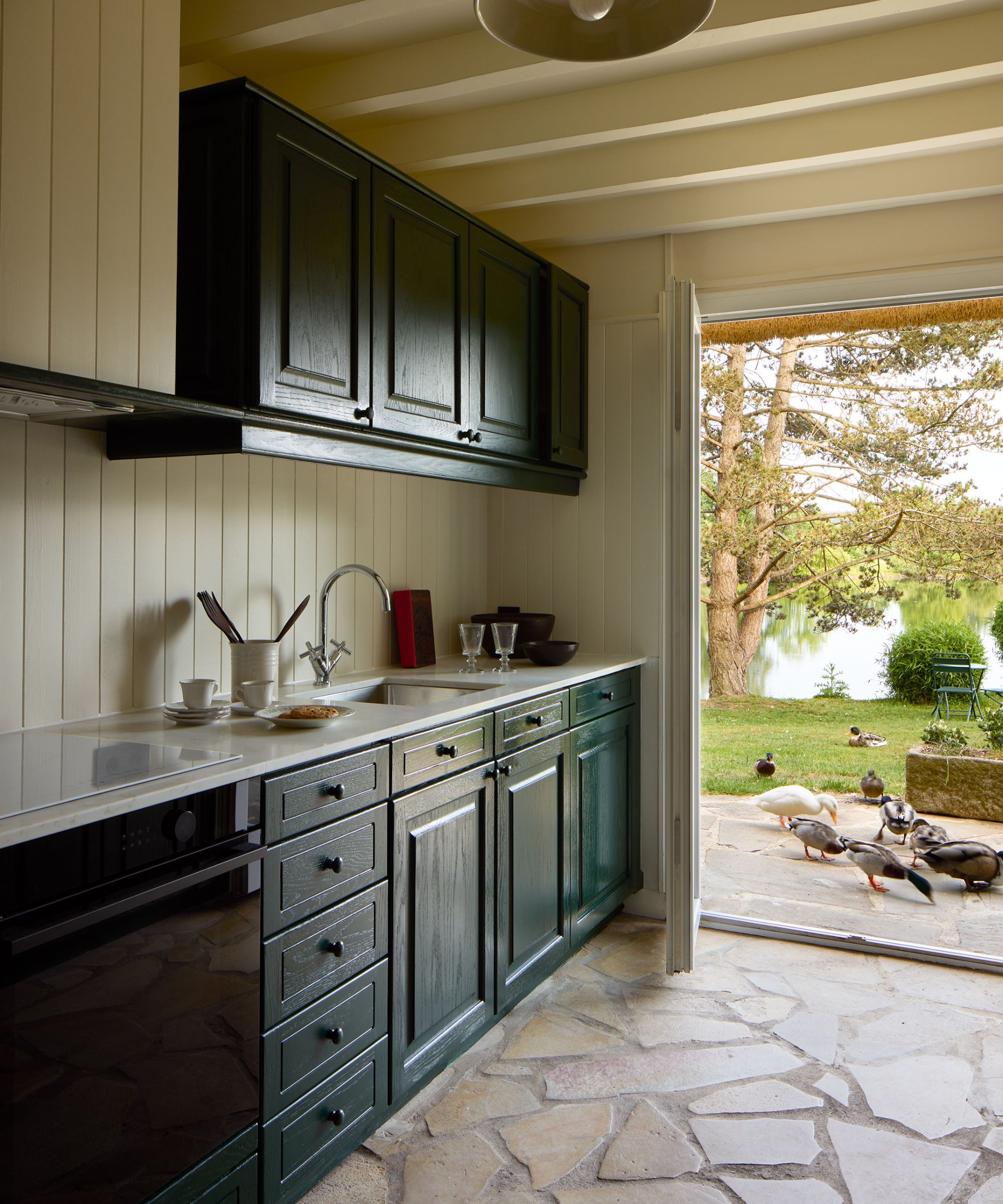 a small deep green wooden kitchen with pale stone floor, ceiling beams and beige painted tongue and groove paneled walls and cooker hood