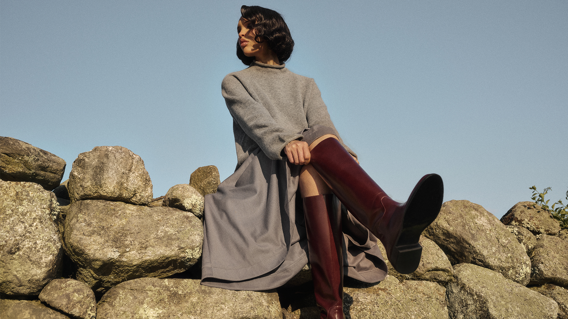 Nara wears gray sweater, gray skirt, and tall red boots while sitting on rocks