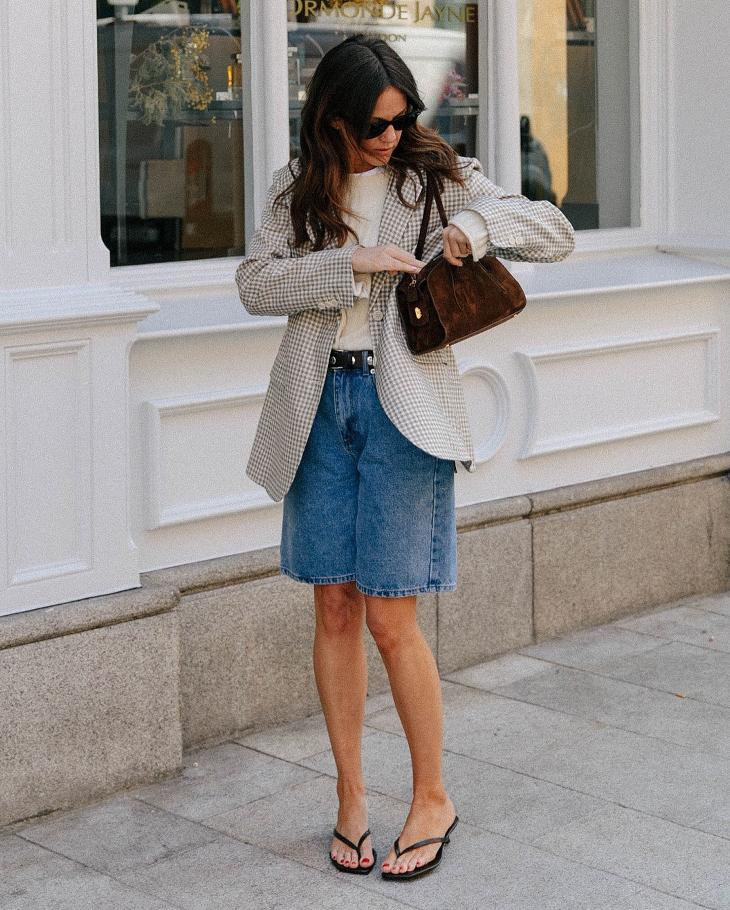 British influencer Kim Turkington on a London sidewalk looking into her brown suede shoulder bag while wearing black sunglasses, a cream sweater, checkered blazer, studded belt, long Bermuda denim shorts, and black flip-flop heeled sandals