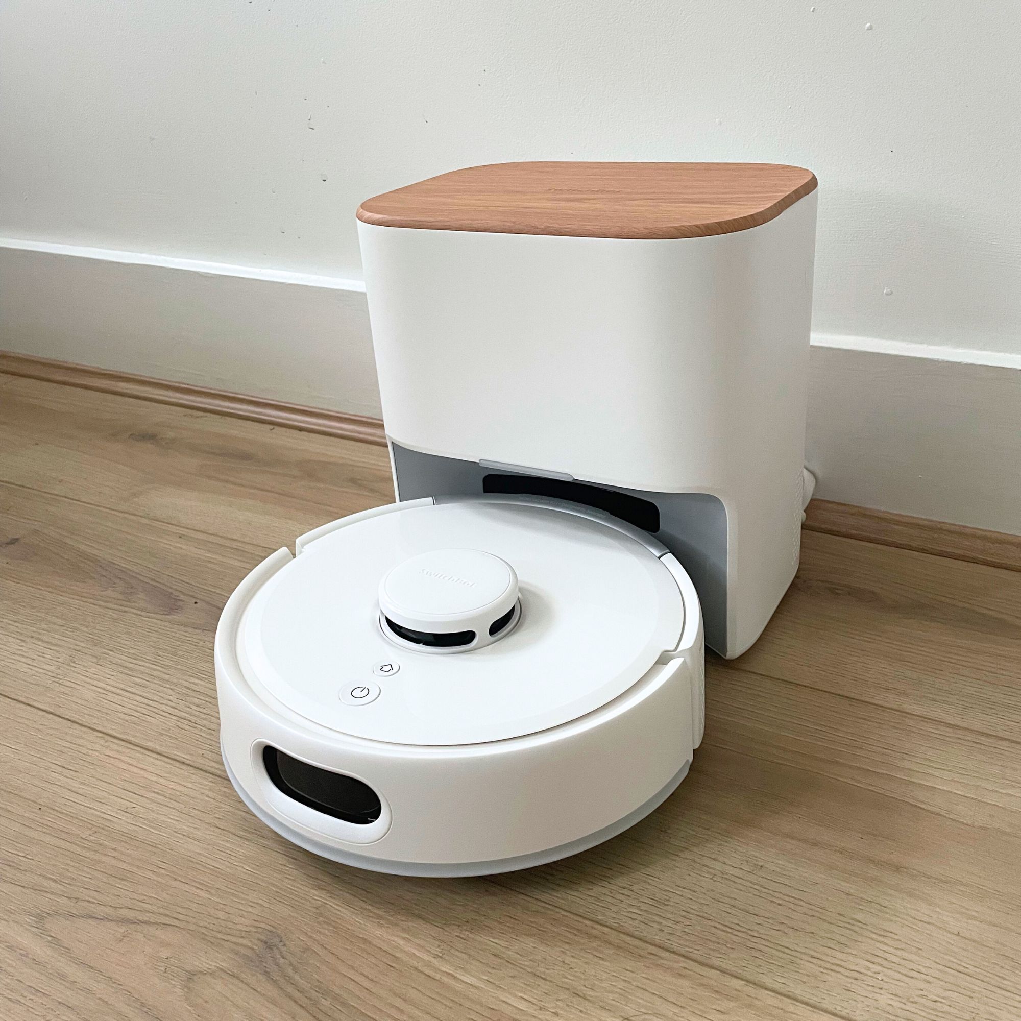 SwitchBot K11+ Robot Vacuum Cleaner with Mop sitting on wood-effect flooring in front of a white wall