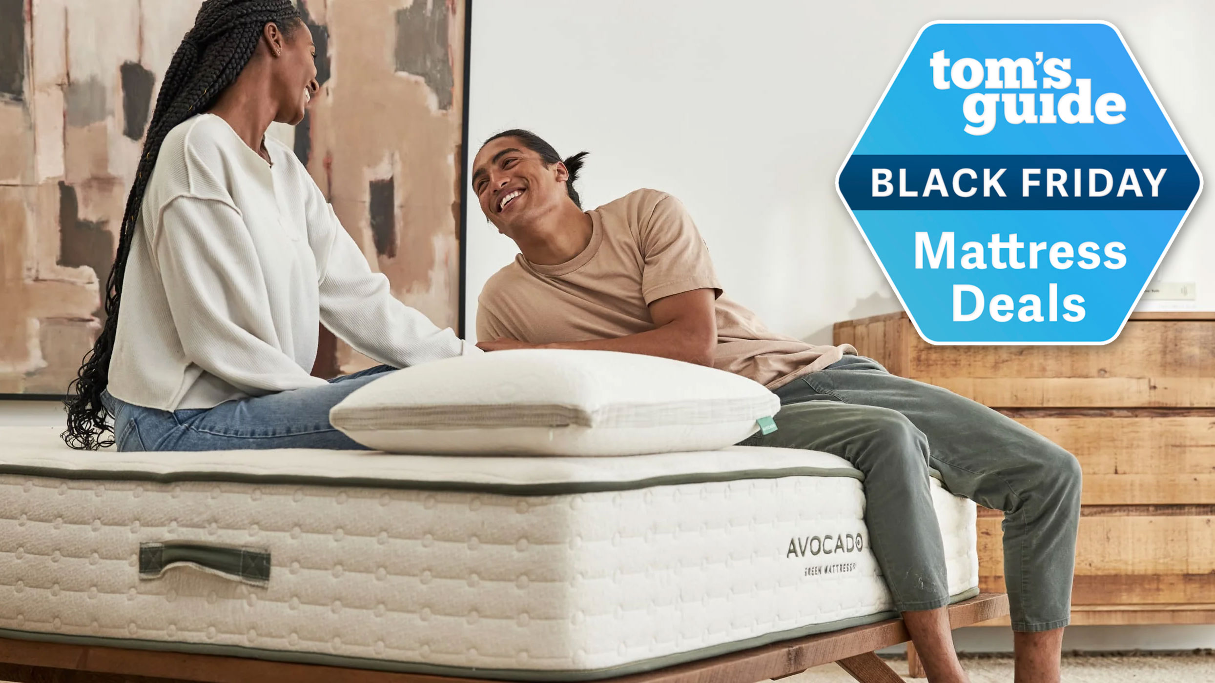 A couple sitting on the Avocado Green Mattress, with a Tom&#039;s Guide Black Friday Mattress Deals badge overlaid