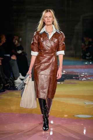 A female model wearing a brown leather dress and knee high leather boots walking down a runway.