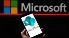 The Microsoft Sharepoint logo, shown on a phone, held in front of the Microsoft logo against a dark background.