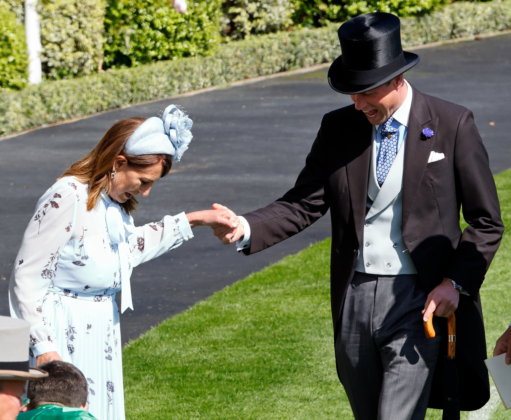 Carole Middleton wearing a blue floral dress and headband holding her hand out to Prince William, dressed in a morning suit and top hat, at Royal Ascot as they laugh