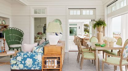 Traditional coastal home with white shiplap walls, large windows, a dining table with green chairs, a floral sofa with a wooden console and two lamps behind it and a green peacock chair in the corner.