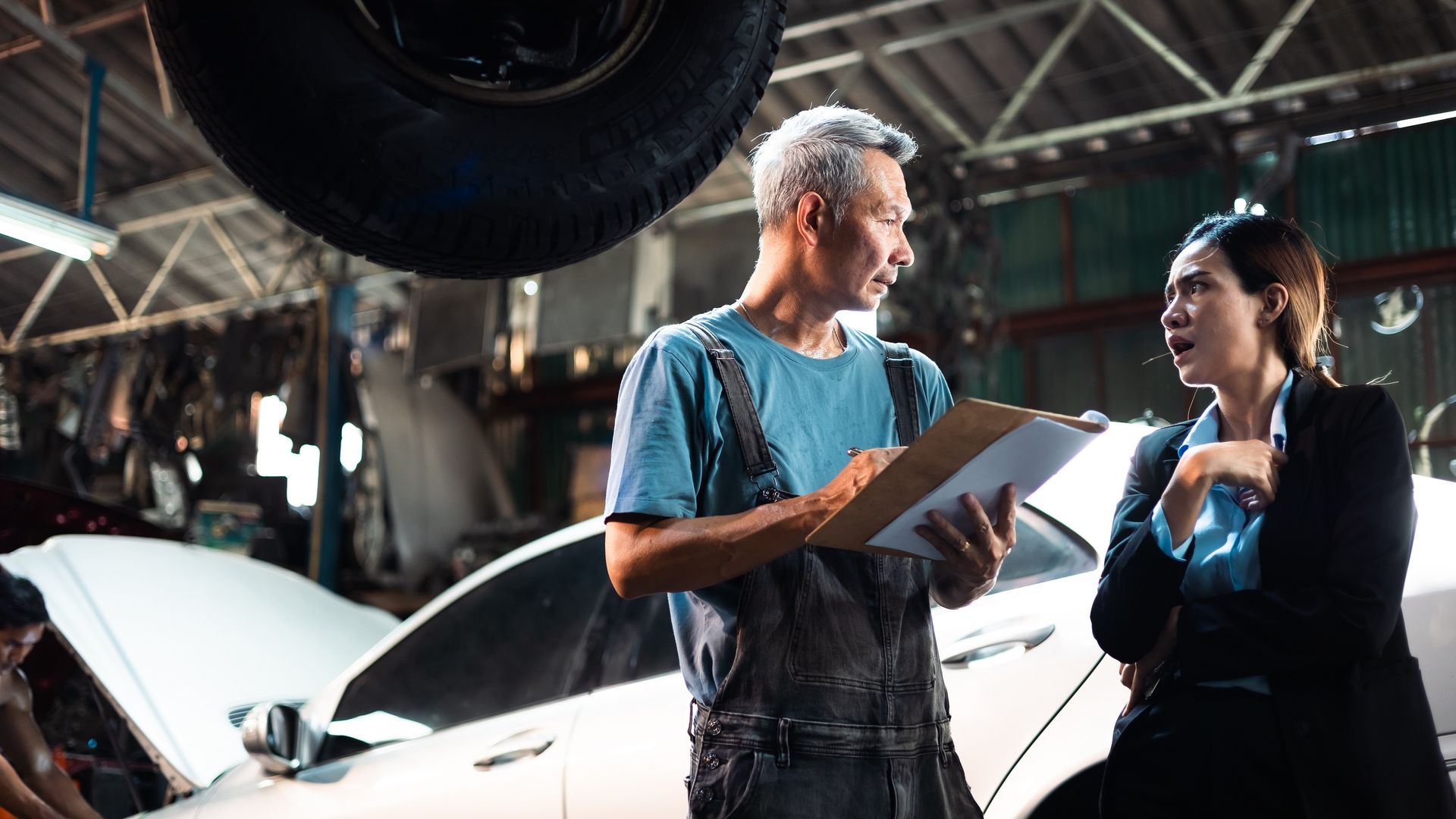 An auto repair worker holding a clipboard talks with an angry customer at a repair shop.