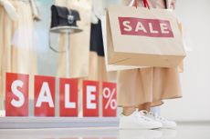 A person holding a shopping bag with the word SALE printed in red, in front of a clothing store window with the word SALE also displayed in red.