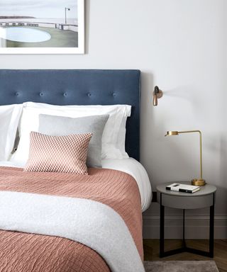 A bed with a blue velvet headboard, pink and white bedding and a simple gold bedside lamp