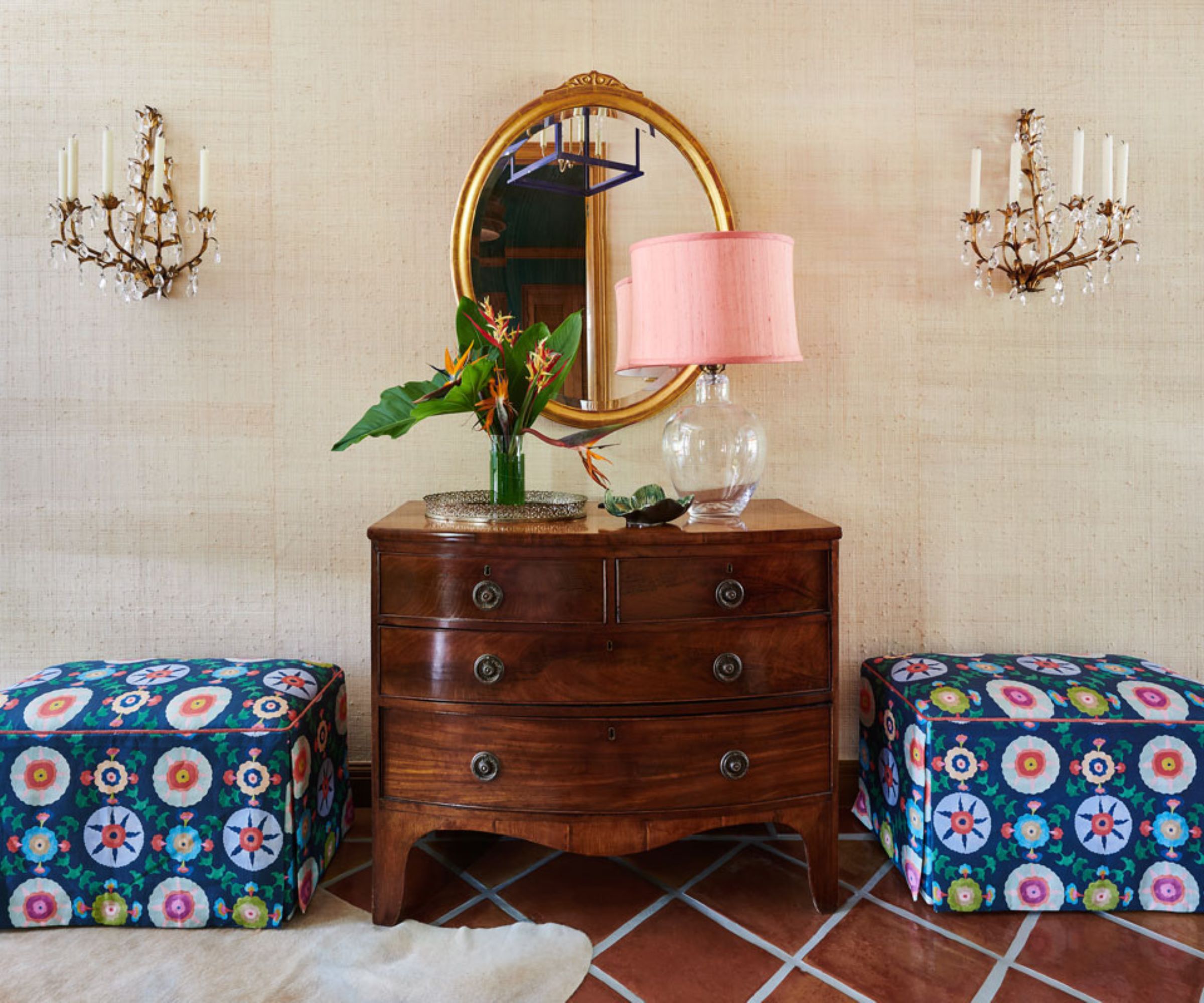 Entryway with tiled floor, sand grasscloth walls, two colourful pouffes beside a mahogany dresser with a large pink lamp on top, a gold round mirror hung above, and two antique candelabras on the wall