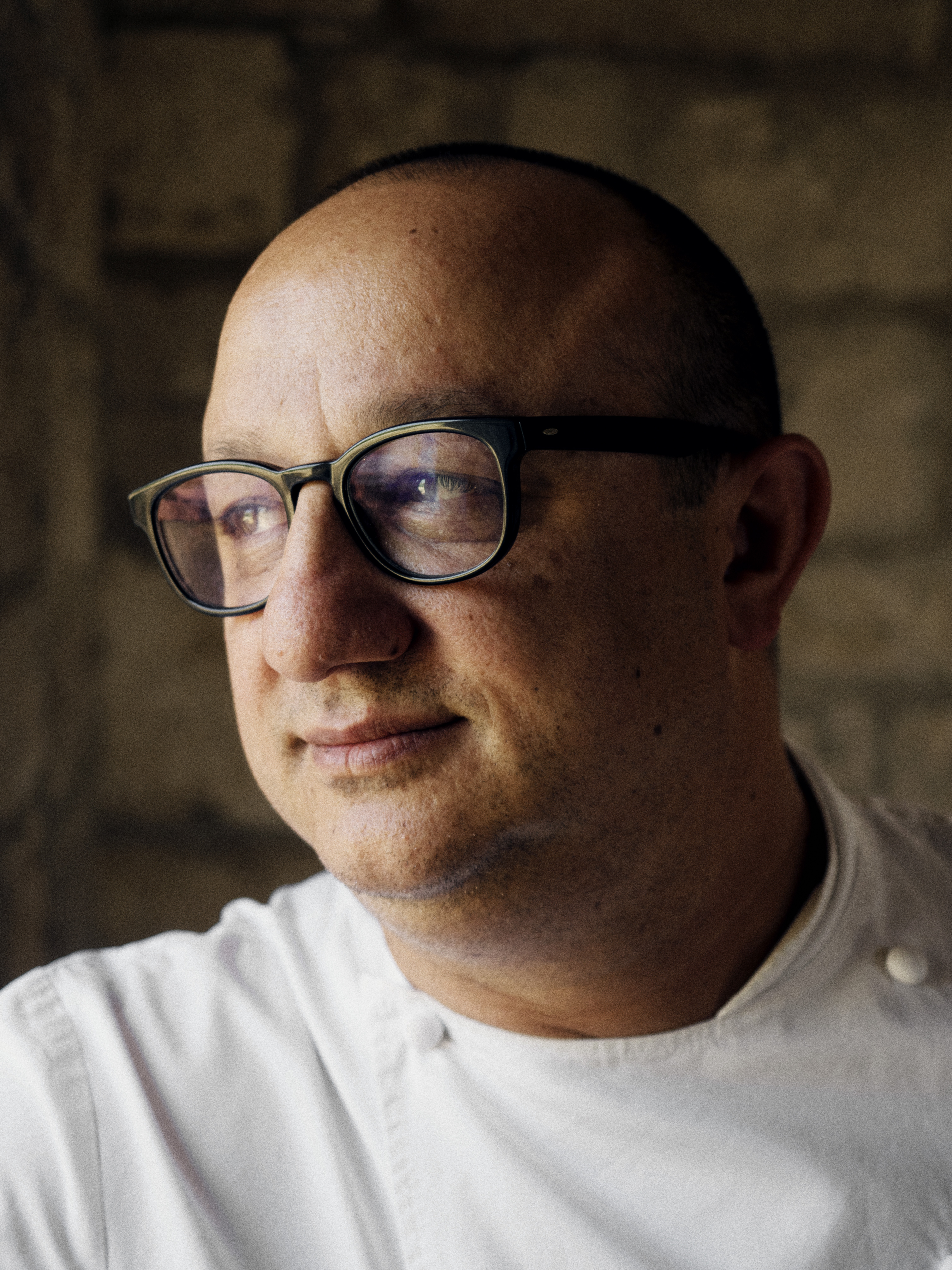 A middle-aged man dressed in a chef shirt glances to his right-hand side while wearing black square glasses in a dimly lit restaurant room.