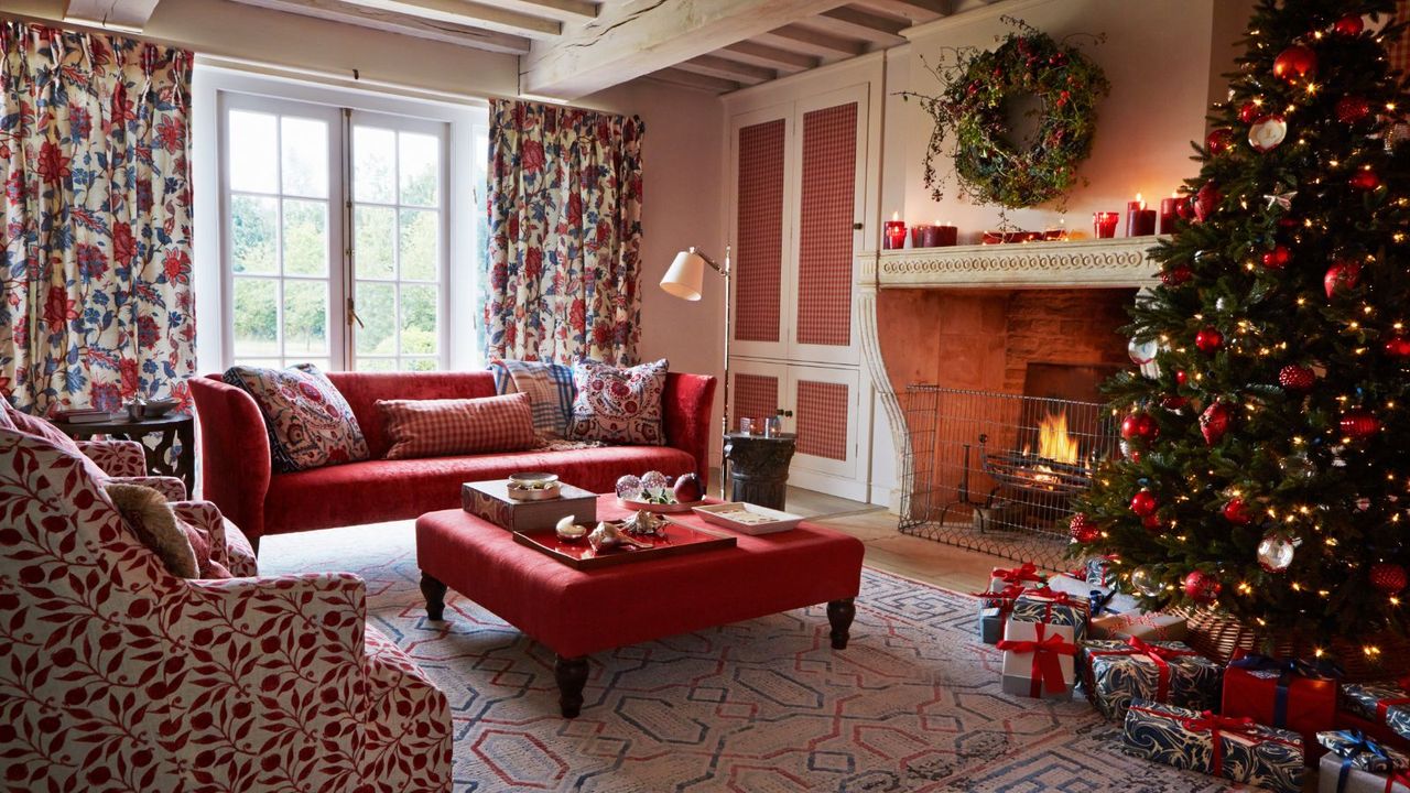 A red festive living room with a large stone fireplace, patterned curtains, a large red couch with a large red ottoman, patterned upholstered chair, patterned rug, and a large decorated christmas tree to the right with wrapped presents beneath. To the left are two large windows and a large cupboard with panelled doors.