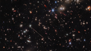 Various warped galaxies are seen as elongated lines, loops, arcs and smudges against the background of space. The JWST's iconic diffraction spikes are seen toward the center-right of the image.