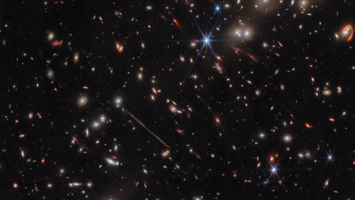 Various warped galaxies are seen as elongated lines, loops, arcs and smudges against the background of space. The JWST&#039;s iconic diffraction spikes are seen toward the center-right of the image.