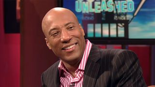 Byron Allen smiles while talking to some comedian guests on Comics Unleashed. He has on a grey suit and pink striped shirt.