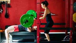 Woman setting up to do a chest press on a rack with supportive personal trainer behind her for help